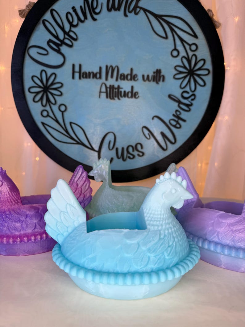 Puede incluir: Un recipiente en forma de gallina azul claro con un dise&ntilde;o detallado, sentado en una base en forma de nido. La imagen tambi&eacute;n muestra otros recipientes en forma de gallina en p&uacute;rpura y verde. Un letrero de madera en el fondo dice "Caffeine and Cuss Words" y "Hand Made with Attitude."
