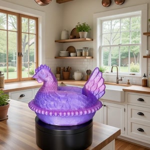 Puede incluir: Un plato decorativo en forma de pollo morado y rosa sobre una base giratoria negra. El plato est&aacute; hecho de un material texturizado y est&aacute; colocado sobre una encimera de madera en un entorno de cocina. La cocina tiene gabinetes blancos y vigas de madera.