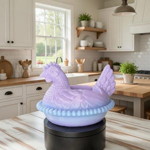 Puede incluir: Un recipiente de cer&aacute;mica en forma de gallina, de color lavanda y azul claro. La gallina se asienta sobre una base redonda azul claro con un borde con volantes. El recipiente est&aacute; sobre un soporte giratorio negro, en una cocina luminosa.