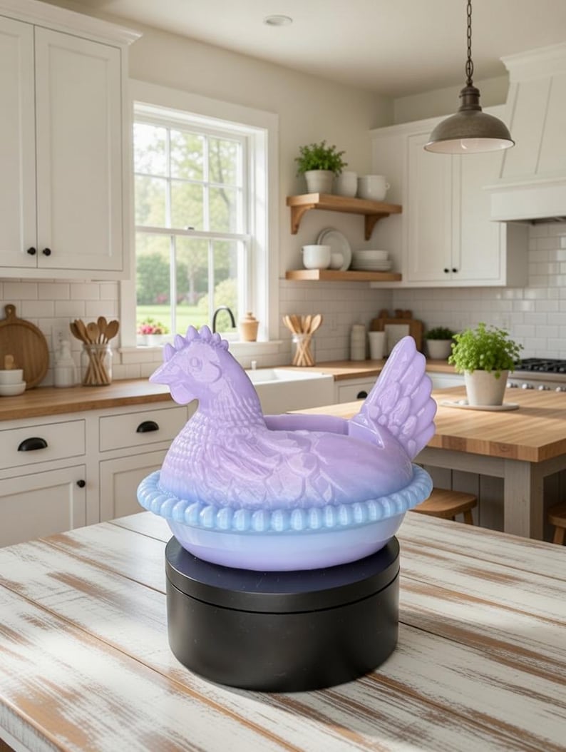 Puede incluir: Un recipiente de cer&aacute;mica en forma de gallina, de color lavanda y azul claro. La gallina se asienta sobre una base redonda azul claro con un borde con volantes. El recipiente est&aacute; sobre un soporte giratorio negro, en una cocina luminosa.