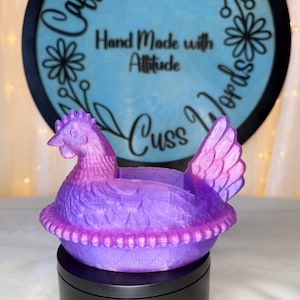 Puede incluir: Un recipiente con forma de pollo morado y rosa. El recipiente est&aacute; sobre una base negra. El fondo presenta un letrero azul con las palabras "Caffeine and Cuss Words" y "Hand Made with Attitude".