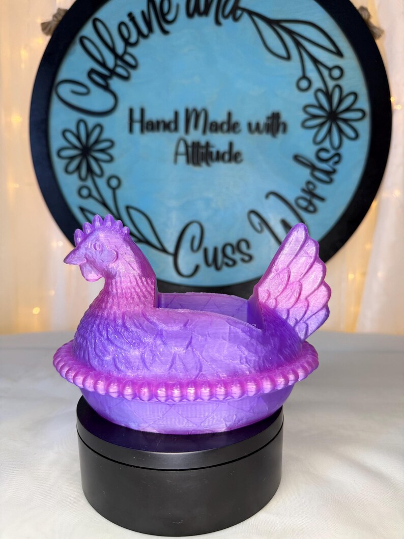 Puede incluir: Un recipiente con forma de pollo morado y rosa. El recipiente est&aacute; sobre una base negra. El fondo presenta un letrero azul con las palabras "Caffeine and Cuss Words" y "Hand Made with Attitude".