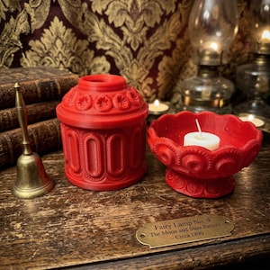 May include: Red decorative candle holder and matching candle dish with a lit candle. The candle holder has a detailed floral design. A brass bell and a sign that reads "Fairy Lamp No. 4 - The Moon and Stars Pattern - Circa 1890" are also visible.