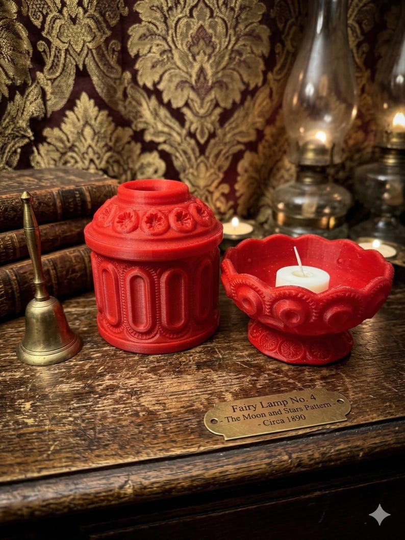 May include: Red decorative candle holder and matching candle dish with a lit candle. The candle holder has a detailed floral design. A brass bell and a sign that reads "Fairy Lamp No. 4 - The Moon and Stars Pattern - Circa 1890" are also visible.