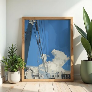Peut inclure: Une impression encadrée représentant un ciel bleu avec des nuages blancs, des lignes électriques et des bâtiments. L'œuvre d'art est dans un cadre en bois clair, évoquant une journée ensoleillée. L'impression est exposée sur un sol en bois.