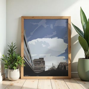 Peut inclure: Une affiche encadrée représentant un paysage urbain sous un ciel nuageux. L'œuvre d'art est de style minimaliste, avec des bâtiments gris et un ciel bleu. Le cadre est en bois clair. Des plantes décoratives sont au premier plan.