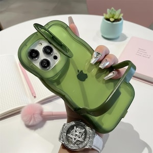May include: A transparent green phone case with a built-in handle and a stand. The case has a wavy design and features the Apple logo. The phone case is designed to protect the phone from damage.