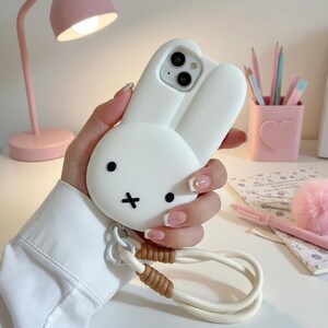 May include: White phone case featuring a 3D bunny design with black eyes and a cross-shaped mouth. The case has a wrist strap with a white cord and brown accents. The phone case is held in a hand.