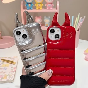 May include: Two phone cases with horn-like accents. One case is metallic silver, the other is bright red. Both cases have a quilted design and a skull and crossbones symbol. The cases are designed to fit a smartphone.