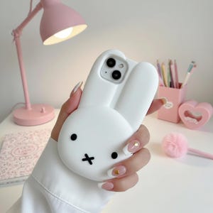 May include: A white phone case shaped like a bunny with black eyes and a cross-shaped mouth. The case has raised ears and a round face. The phone case is held in a hand with manicured nails. A pink desk lamp and other pink desk accessories are in the background.