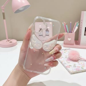 Könnte beinhalten: Transparente Handyhülle mit schillerndem Engelsflügel-Design. Die Hülle hat einen herzförmigen Ausschnitt oben und kleine sternförmige Glitzerakzente. Die Flügel sind weiß mit einem perlmuttartigen Schimmer.