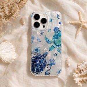May include: A clear smartphone case featuring a printed design of two sea turtles in shades of blue and teal, surrounded by small seashells and starfish illustrations. The case has a cutout for a triple-lens camera system on the top left corner.