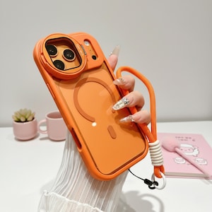 May include: Orange phone case with a camera lens protector and a matching orange lanyard. The case has a clear back with a circular design. The lanyard is attached with a white and black clasp.