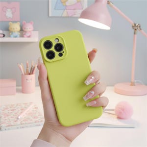 May include: A lime green phone case with a camera module featuring four lenses. The case is held in a hand with long, decorated fingernails. A pink desk lamp and decorative items are in the background.