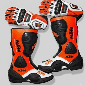 Könnte beinhalten: Ein Paar orangefarbene, schwarze und weiße KTM Racing Team Motorradstiefel und passende Handschuhe. Die Stiefel und Handschuhe tragen das Red Bull Logo und das KTM-Branding. Die Stiefel sind hoch, mit schwarzer Sohle und orangefarbenem Obermaterial.