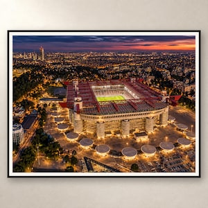 May include: An aerial view of the San Siro stadium in Milan at dusk, featuring its distinctive red structural supports and cylindrical towers. The illuminated stadium sits amidst a sprawling urban landscape under a vibrant sunset sky.