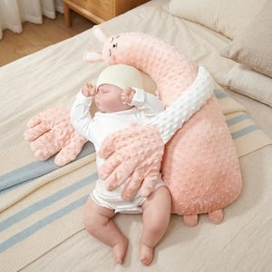 Könnte beinhalten: Ein schlafendes Baby in einem weißen Strampler und einer hellgelben Mütze wird von einem rosa Plüschkissen in Hasenform mit großen Händen umarmt. Das Kissen hat eine strukturierte Oberfläche und das Wort "Love" ist darauf gestickt.