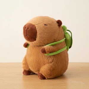 Op de afbeelding: Een bruin pluche capybaraspeelgoed met een donkerbruine neus en oren. Het heeft een kleine groene rugzak met groene bandjes. Het speelgoed staat op een houten oppervlak.