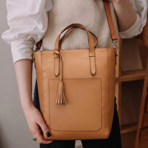 May include: A tan vertical tote bag featuring two top handles, a detachable shoulder strap with silver-toned metal hardware, and a front slip pocket. A decorative leather tassel hangs from one of the handle attachments on the front exterior.