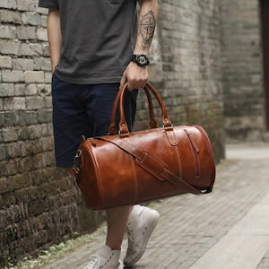 May include: A cylindrical brown leather duffel bag featuring two top handles, a detachable shoulder strap with a padded section, and a side zipper pocket. The bag has visible stitching details and metal hardware connecting the straps to the body.