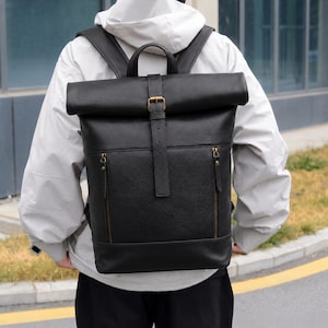 Puede incluir: Una mochila negra con cierre enrollable, superficie texturizada, una correa vertical central con hebilla metálica en tono latón y dos bolsillos frontales con cremallera y herrajes a juego. Incluye un asa superior y dos correas para los hombros.