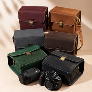 Puede incluir: Seis bolsas rectangulares para cámara en cuero burdeos, tostado, gris, marrón oscuro, verde y negro, cada una con cierre de hebilla metálica en tono latón, costuras blancas en contraste y anillas en D. Un cuerpo de cámara negro y un objetivo están delante.