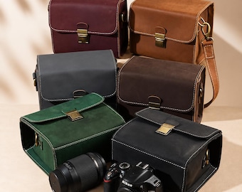 May include: Six rectangular camera bags in burgundy, tan, gray, dark brown, green, and black leather, each featuring a brass-toned metal clasp closure, white contrast stitching, and side D-rings. A black camera body and a lens are placed in front.