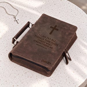 May include: A brown leather Bible cover featuring a handle, a zipper closure, and an engraved cross. The text reads: I CAN DO ALL THINGS THROUGH CHRIST WHO STRENGTHENS ME. PHILIPPIANS 4:13. The name DAVID is engraved below. A silver cross necklace is nearby.