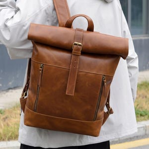 Peut inclure: Un sac à dos en cuir marron clair à fermeture par enroulement, doté d'une sangle verticale centrale avec boucle en métal, d'une poignée supérieure et de deux poches zippées verticales sur le devant. Il possède des bretelles réglables et des ferrures en laiton.