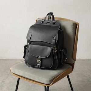 May include: A black leather backpack with a flap closure, two front pockets, and side pockets. The backpack has gold-colored buckles and white stitching. It is sitting on a gray chair.
