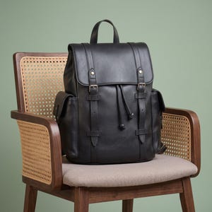 May include: A black backpack featuring a top handle, a fold-over flap with two buckle straps, and a drawstring closure. The bag includes two side pockets and vertical strap detailing on the front, constructed with a smooth, matte finish.