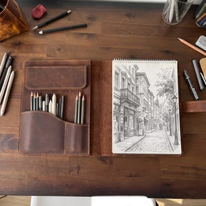 May include: A brown leather sketchbook cover with an integrated organizer featuring multiple slots for pencils and a spiral-bound sketchbook displaying a detailed graphite drawing of a European street scene with buildings and street lamps.