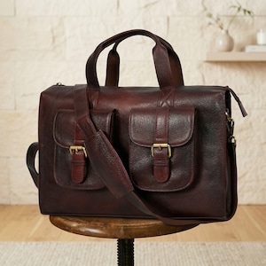 May include: A dark brown leather briefcase with two front pockets secured by brass buckles. The bag has a top handle and a detachable shoulder strap. The briefcase is sitting on a wooden stool.