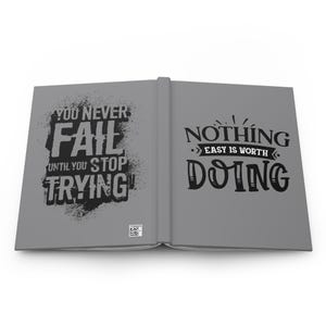 Pode incluir: Um diário cinza com citações motivacionais pretas. A página esquerda diz "You Never Fail Until You Stop Trying." A página direita diz "Nothing Easy Is Worth Doing."
