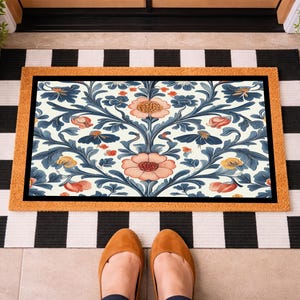 May include: A rectangular doormat featuring a central panel with a symmetrical floral pattern in shades of peach, blue, and yellow on a cream background, framed by a textured tan border, layered over a black and white striped rectangular mat.