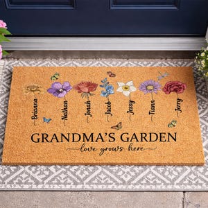 May include: A rectangular doormat with a light brown background features a floral design with names. The words "GRANDMA'S GARDEN" and "love grows here" are printed in black. Colorful flowers and butterflies are also depicted.