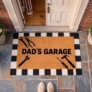 May include: A rectangular doormat with the text "DAD'S GARAGE" and tool illustrations. The mat is bordered with black and white squares and is made of a natural fiber material. The tools include a wrench, pliers, screwdriver, and hammer.