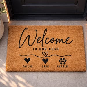 May include: A rectangular coir doormat with the words "Welcome to our home" in black script. Below are three icons: a heart, a heart, and a paw print, each with a name: Taylor, John, and Charlie.