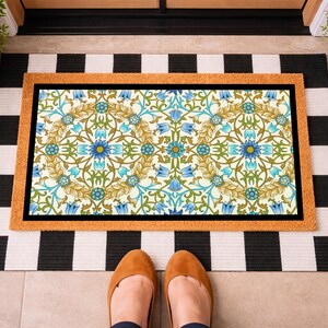 May include: A rectangular doormat featuring a central panel with a repeating floral pattern in shades of blue, green, and gold, framed by a tan border. The mat is layered over a larger rectangular rug with a black and white horizontal stripe pattern.