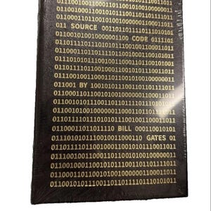 May include: A rectangular black book wrapped in clear plastic. The cover features gold text consisting of binary code sequences interspersed with the words SOURCE CODE BY BILL GATES, arranged in horizontal lines across the entire front surface.