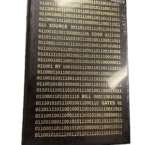 May include: A rectangular black book wrapped in clear plastic, featuring gold binary code printed on the cover. The text "SOURCE CODE BY BILL GATES" is integrated into the binary pattern, with the words appearing in a horizontal line.