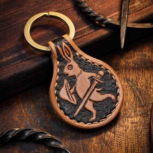 May include: Leather keychain featuring a brown rabbit warrior with a sword and shield on a black background. The keychain has a gold-colored ring and a brown border. The background is a dark wood surface.