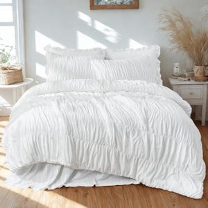 May include: A white bed set with a textured, ruched design. The set includes a comforter, two pillow shams, and a bed skirt. The bedding is arranged on a wooden floor, with a white nightstand and window in the background.