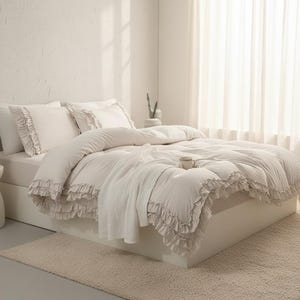 Könnte beinhalten: Ein cremefarbenes Bettwäsche-Set mit Bettdecke, Kissen und einer Decke. Die Bettdecke und Kissenbezüge haben Rüschenkanten. Ein kleiner Teppich und eine Tasse sind ebenfalls zu sehen, was eine gemütliche Schlafzimmerszene schafft.