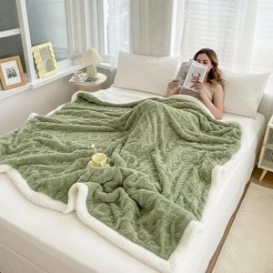 Può includere: Una coperta verde salvia con un motivo strutturato e un bordo in sherpa bianco è drappeggiata su un letto. Una persona legge un libro sotto la coperta. Una tazza gialla con un cucchiaio è sulla coperta.
