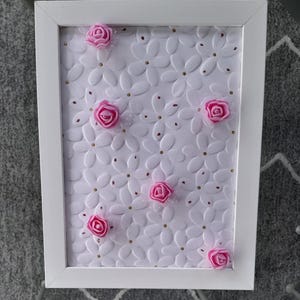 May include: A white framed decorative piece featuring a white floral pattern with raised details. Pink fabric roses are affixed to the surface, interspersed with small gold-colored accents. The frame is square.