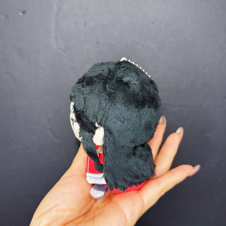 Pu&ograve; includere: Una piccola bambola di peluche con capelli di pelliccia neri, un vestito rosso e un viso bianco. La bambola ha una catena d'argento attaccata in alto. La bambola &egrave; tenuta in una mano su uno sfondo scuro.