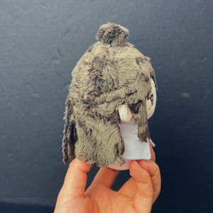 Pu&ograve; includere: Una bambola di peluche con una copertura per la testa strutturata in finta pelliccia marrone e un vestito bianco. La bambola ha un piccolo viso rotondo con tratti dipinti. La bambola &egrave; tenuta in mano da una persona.