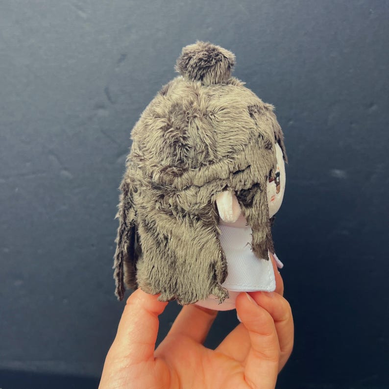 Pu&ograve; includere: Una bambola di peluche con una copertura per la testa strutturata in finta pelliccia marrone e un vestito bianco. La bambola ha un piccolo viso rotondo con tratti dipinti. La bambola &egrave; tenuta in mano da una persona.