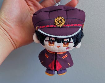 Può includere: Un portachiavi in peluche raffigurante un personaggio dai capelli neri con tratti del viso ricamati, che indossa un berretto militare viola scuro con un emblema dorato e una giacca abbinata con dettagli rossi, appeso a una catenella in metallo argentato.